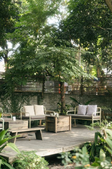 A cozy outdoor garden area featuring a wooden deck with three light-colored seated lounge chairs, some with striped cushions, arranged around a central wooden table. The deck is bordered by lush, gree