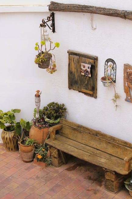 A cozy outdoor garden area featuring a wooden deck with three light-colored seated lounge chairs, some with striped cushions, arranged around a central wooden table. The deck is bordered by lush, gree