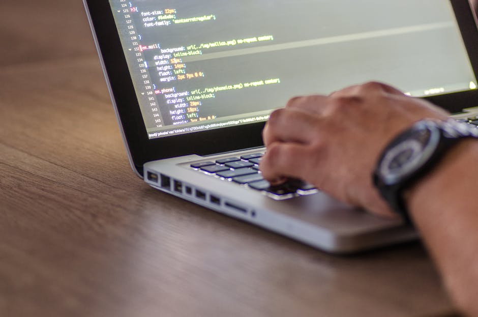 A person's hand is seen typing on the keyboard of a silver laptop placed on a wooden desk. The screen displays lines of colorful programming code, suggesting the individual is engaged in coding or web